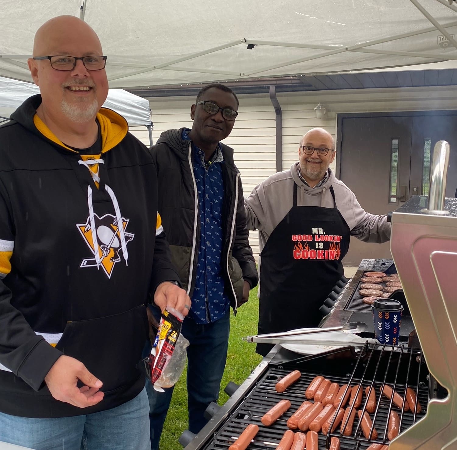 Three men standing around a grill with hot dogs on it.