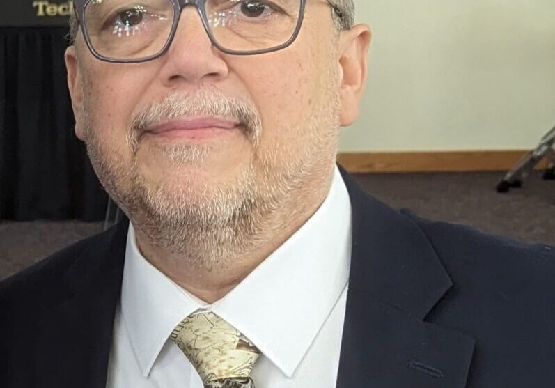 A man in glasses and a suit standing next to drums.