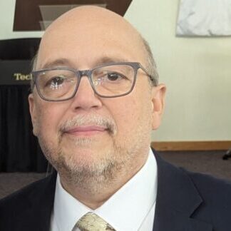 A man in glasses and a suit standing next to drums.